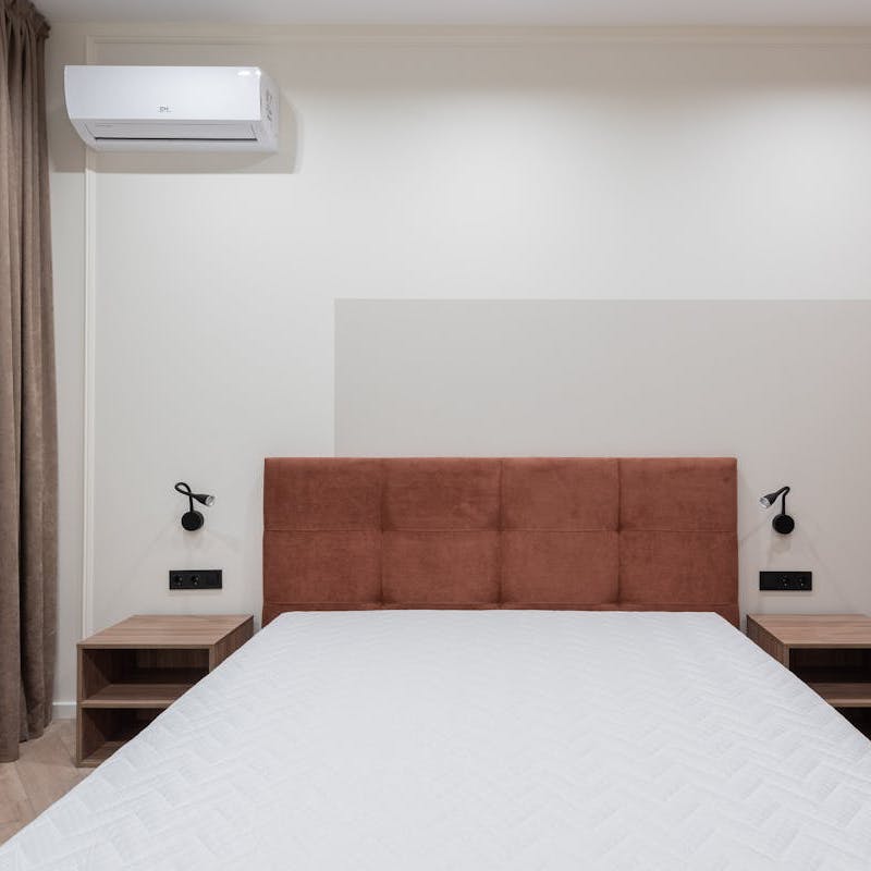 An air conditioner unit on the wall of a bedroom, which is entirely white except for the brown headboard of the bed.